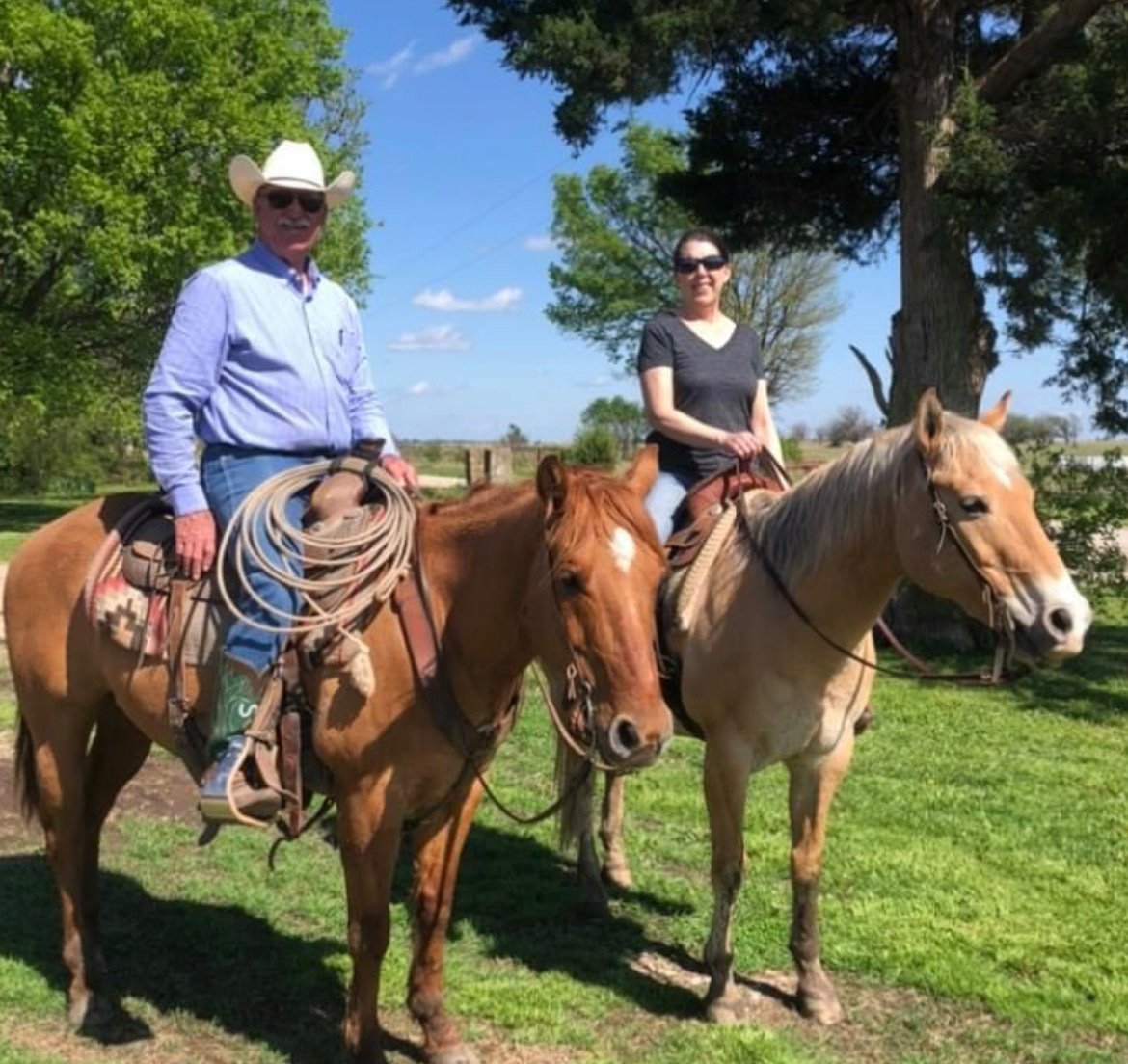 Two people riding horses in a grassy park area with trees and a blue sky in the background. The man is wearing a cowboy hat, sunglasses, and a light blue shirt with a lasso on his saddle. The woman is wearing sunglasses and a black shirt.